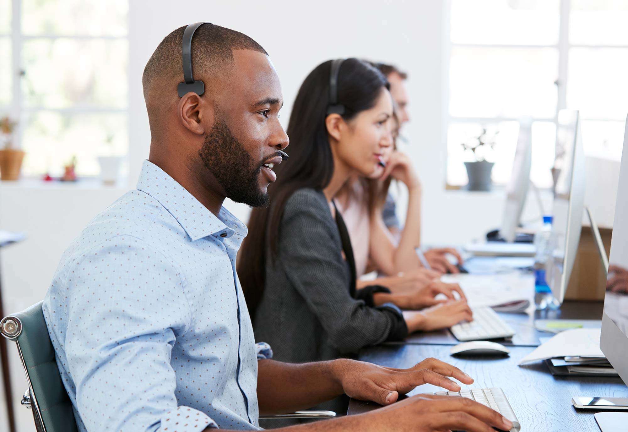 Men and women sit side by side in an office working from computers with telephone headsets on