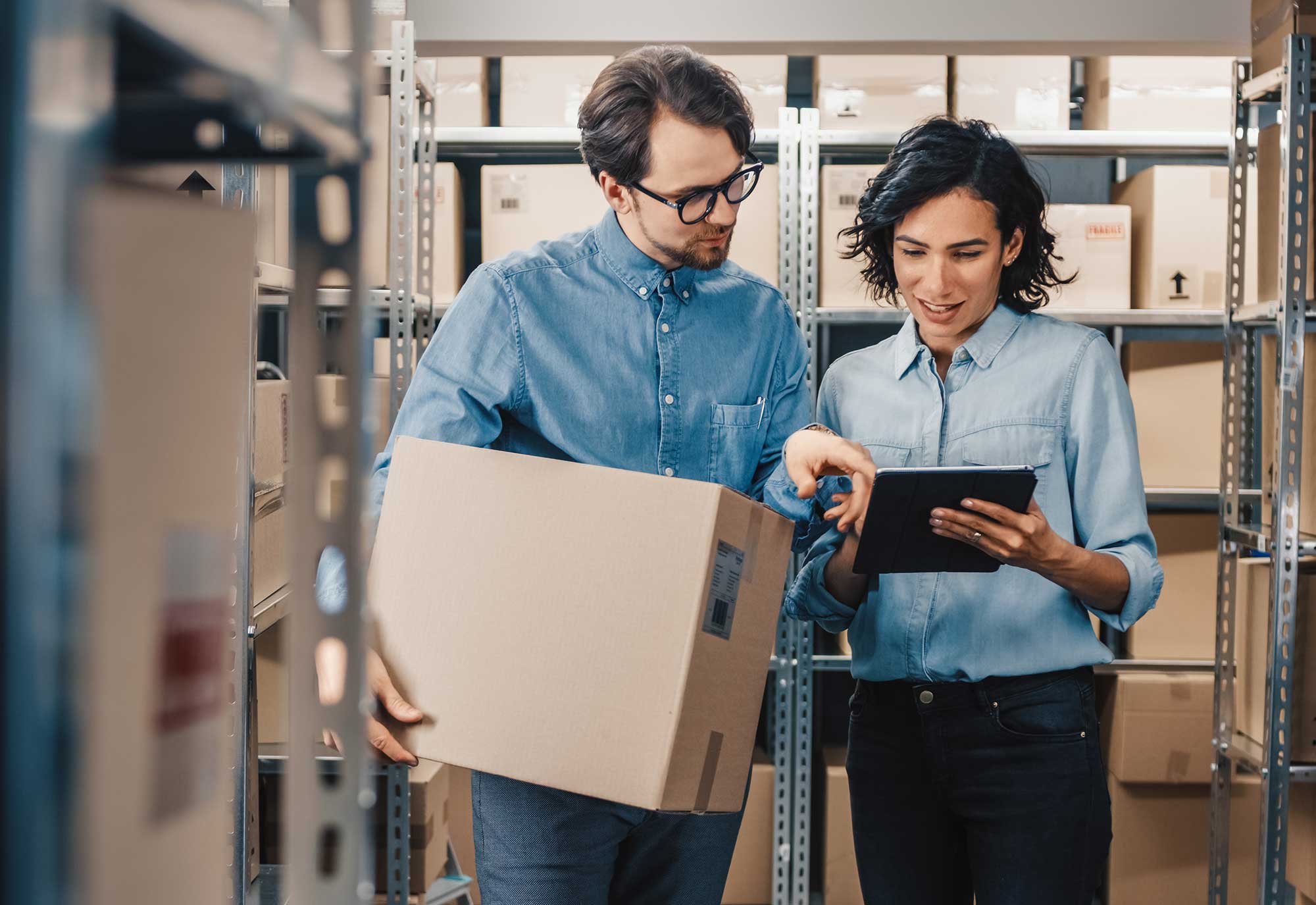 Man hold a large cardboard box while a woman checks information on an tablet device in a retail distribution centre