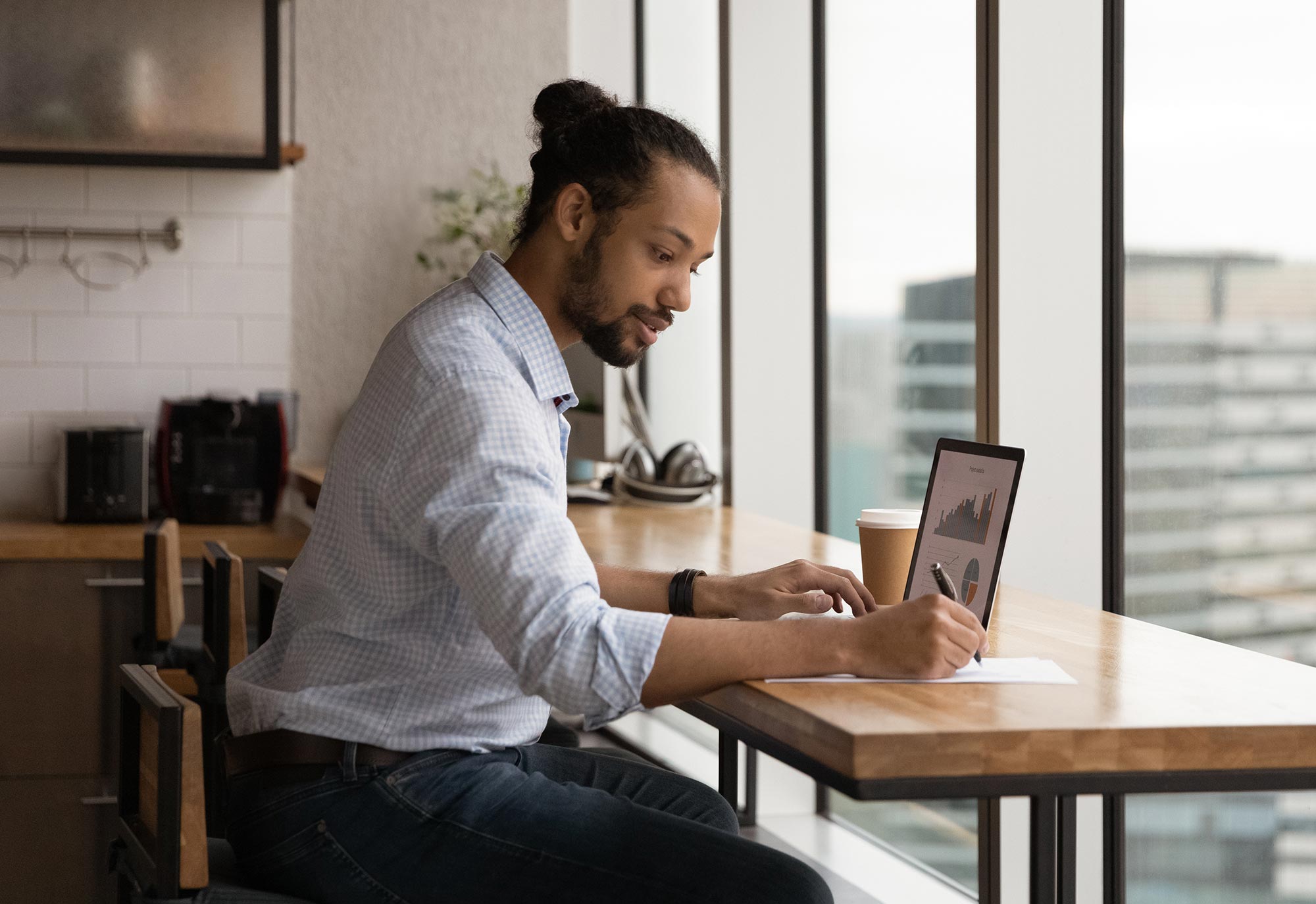 young man sits at a table in a cafe with laptop open and making notes on paper
