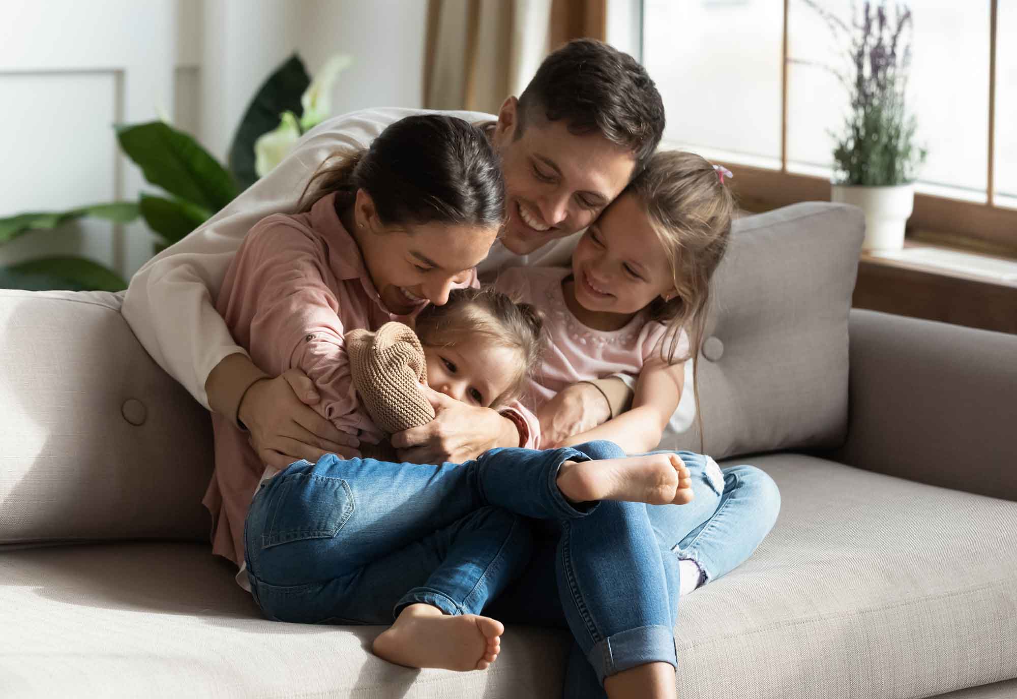 Man cuddles woman, child and baby as they sit and play on a sofa in their living room